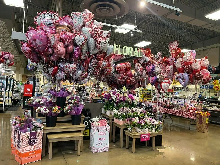 A supermarket floral section adorned with a large display of balloons and flowers, highlighting consumerism and price differences.