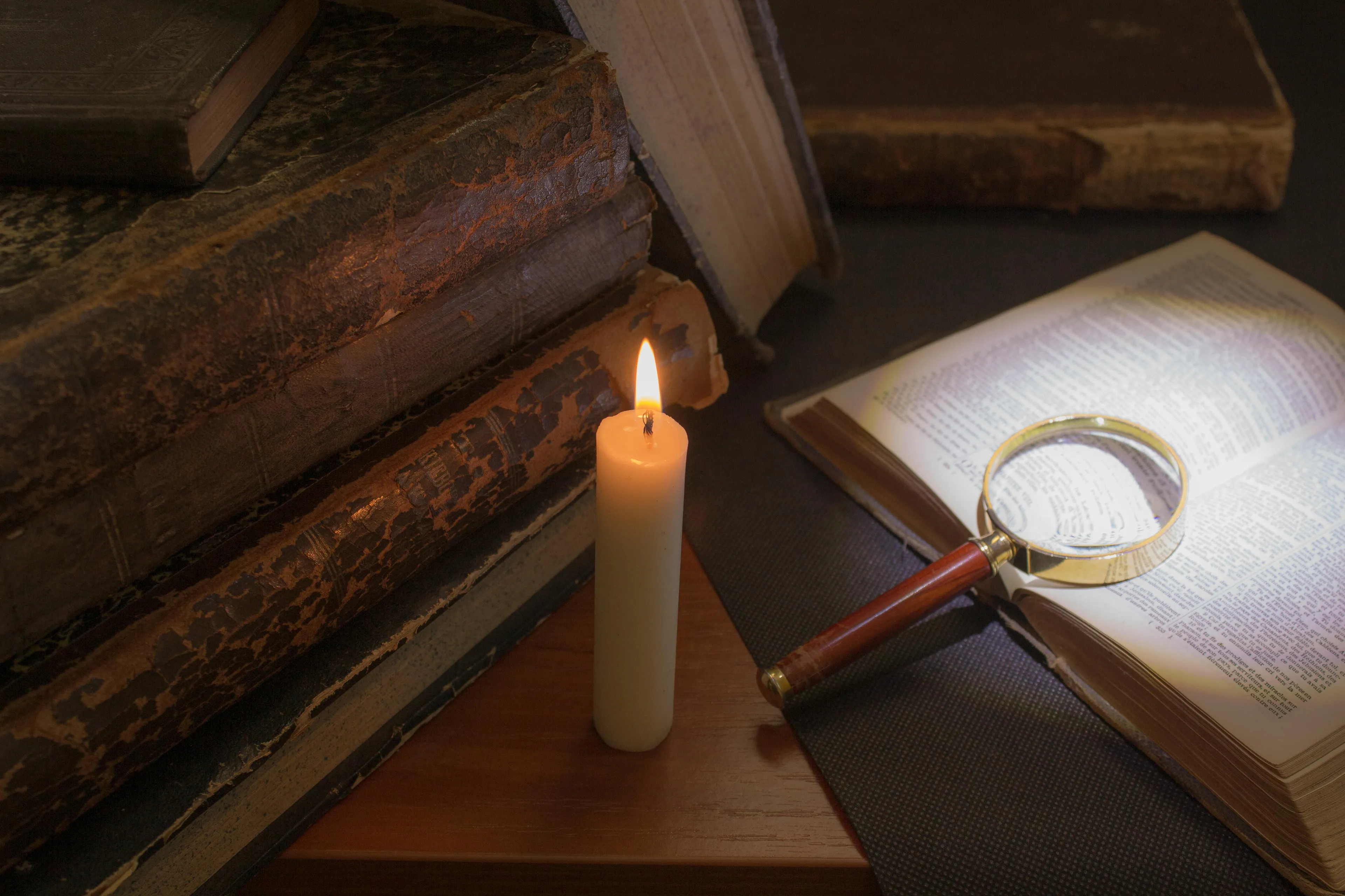 Stack of old books and open book with magnifying glass beside lit candle symbolizing philosophy and nature of knowledge study.