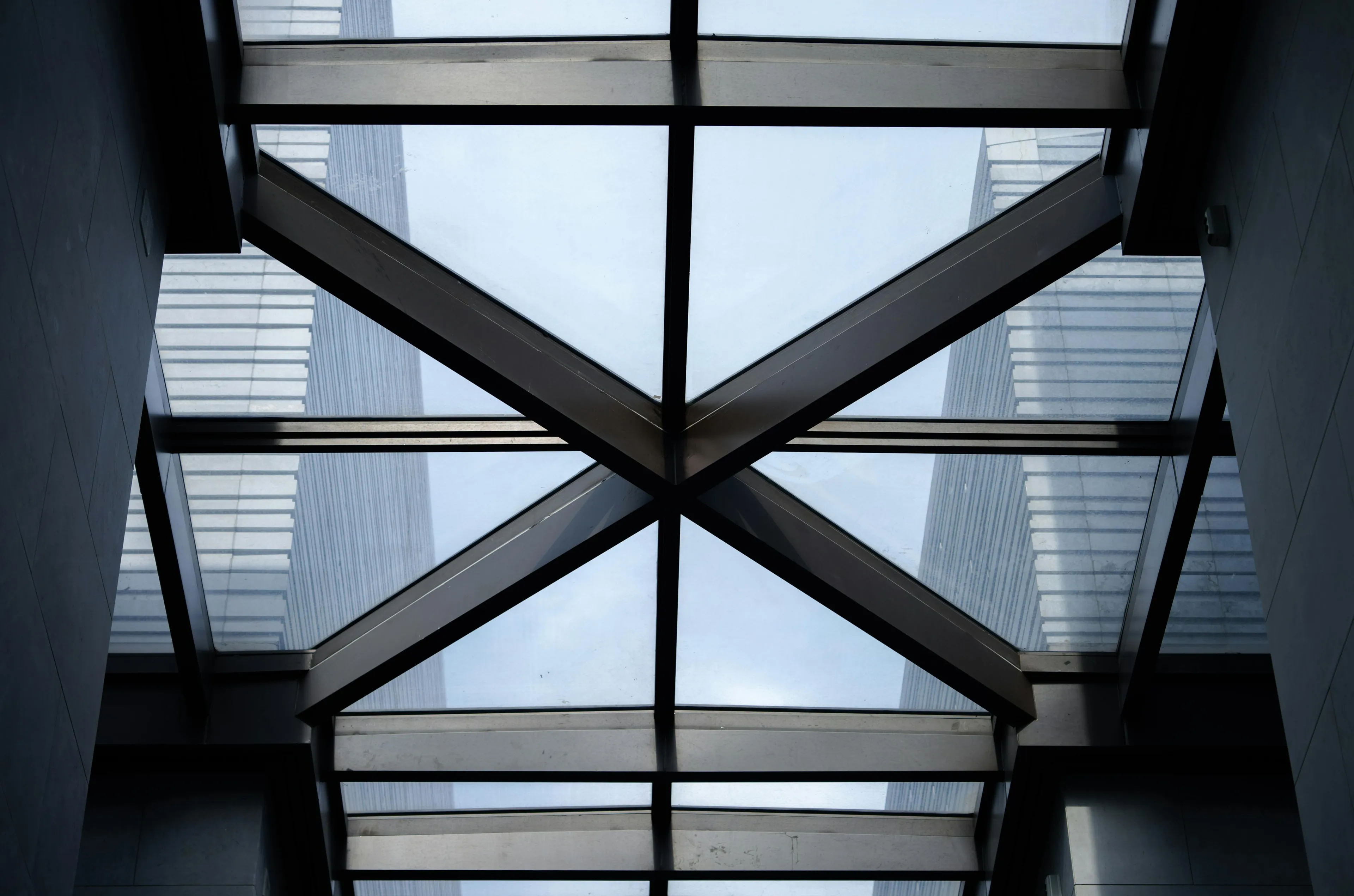 Steel beams forming a cantilever structure supported at the base with glass panels and building reflections overhead.