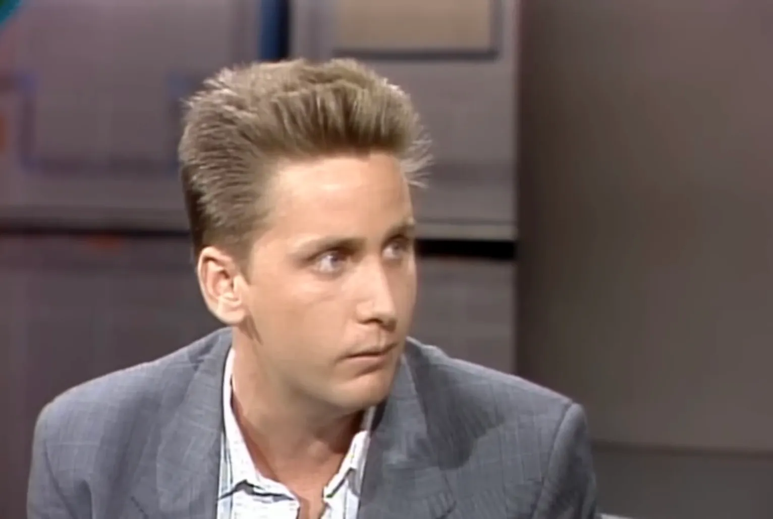 Young 80s celeb, key member of the Brat Pack, wearing a gray blazer during a television interview.