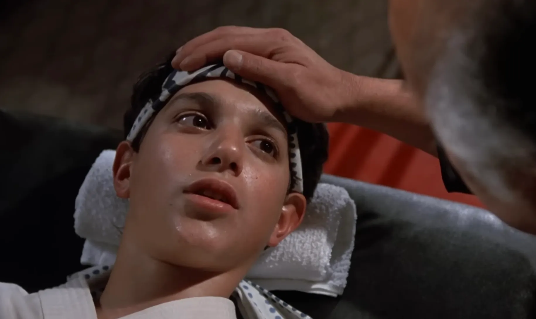 Teen actor from the 80s receiving care with a hand on his forehead, wearing a karate headband and gi.