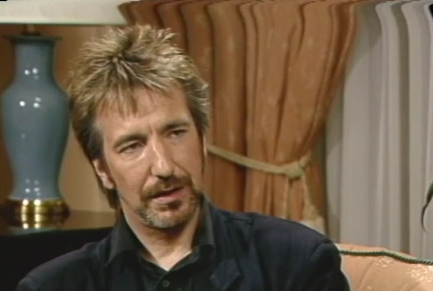 Actor from 80s known for playing an unforgettable villain, seated indoors with a serious expression and casual attire.