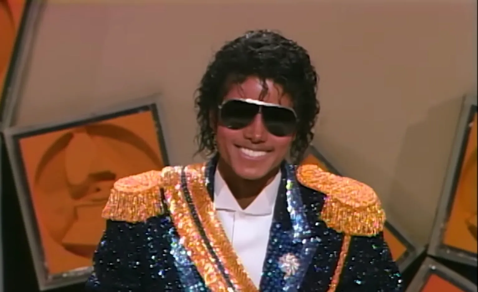 80s celeb wearing a sparkling military-style jacket and sunglasses smiling at an award show backdrop.