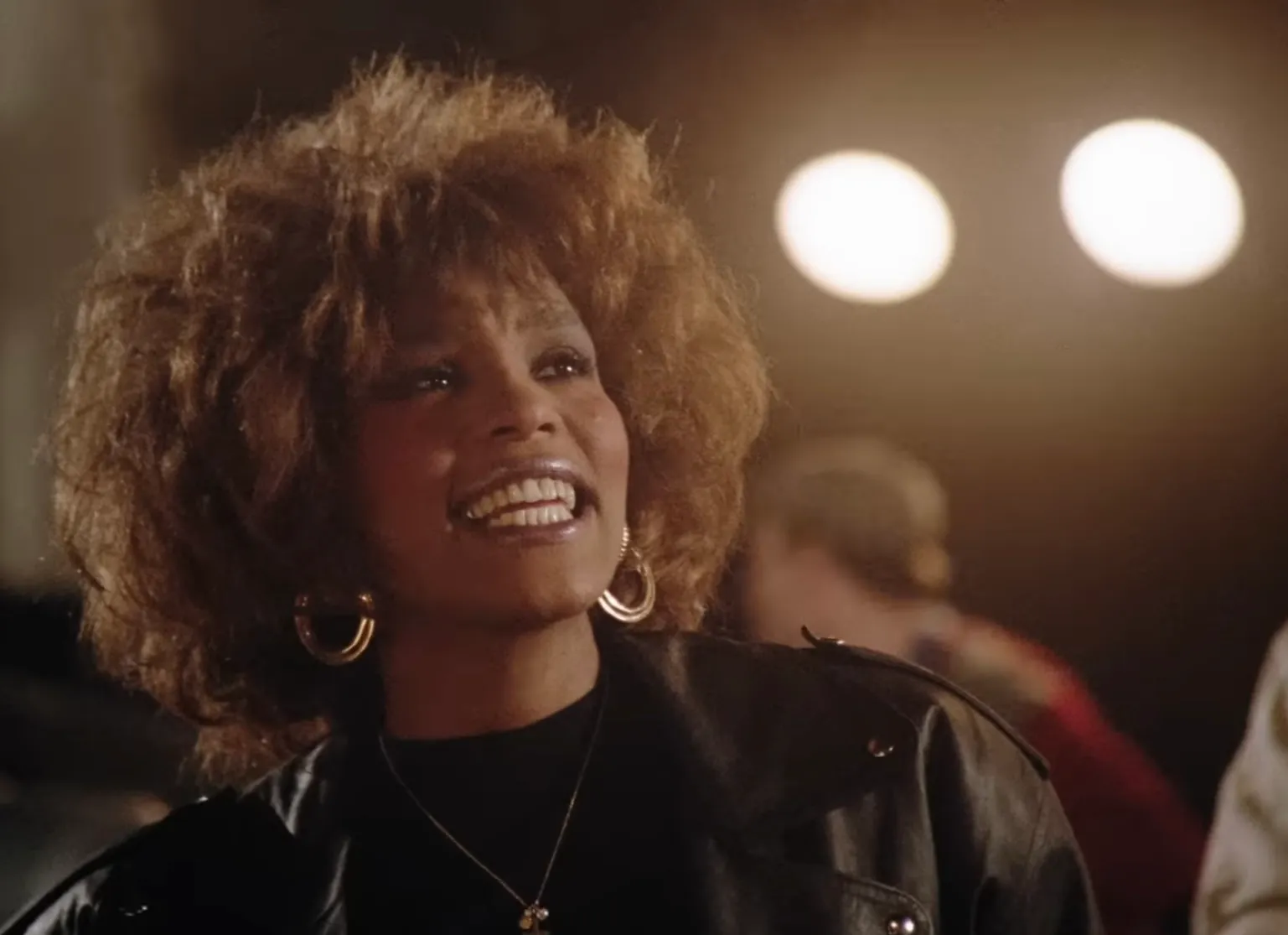 80s celeb with a voice that defined the decade, smiling with big hair, hoop earrings, and a leather jacket under bright lights.