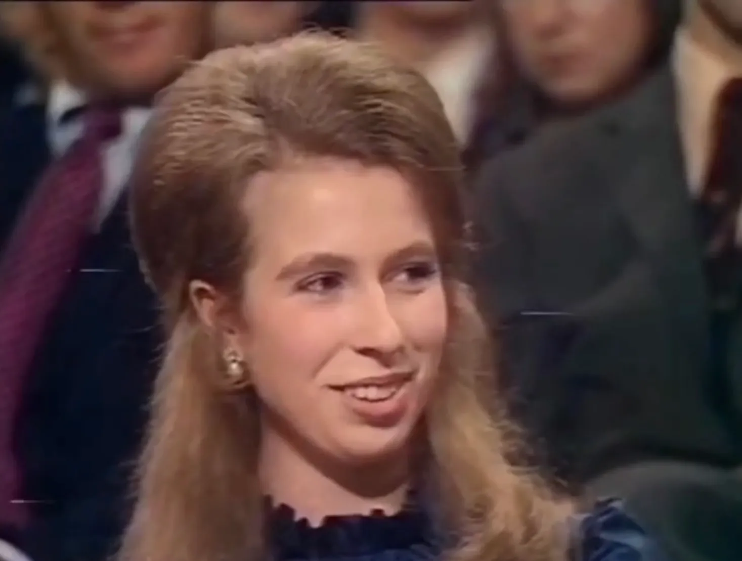 Princess Anne smiling in 80s fashion style with voluminous hair and pearl earrings at an indoor event.