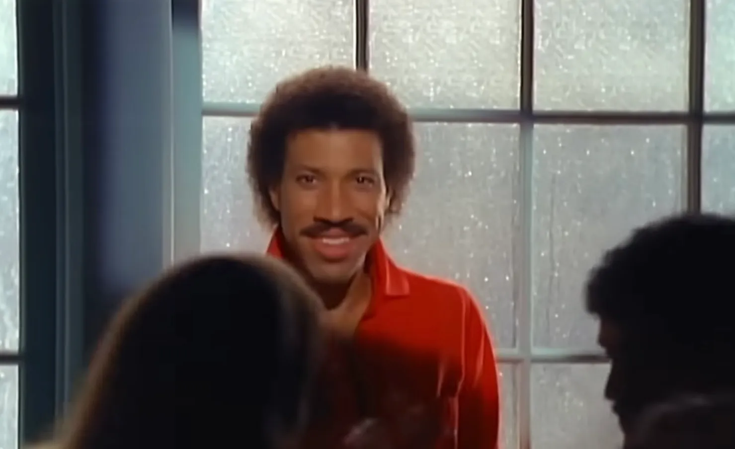 1980s celeb with smooth voice and hit All Night Long smiling indoors with frosted window behind him.