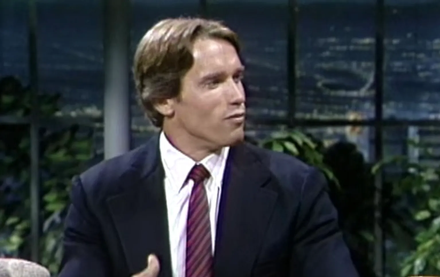 Young 80s action hero in a suit and tie appearing on a late night talk show set with city lights in background.