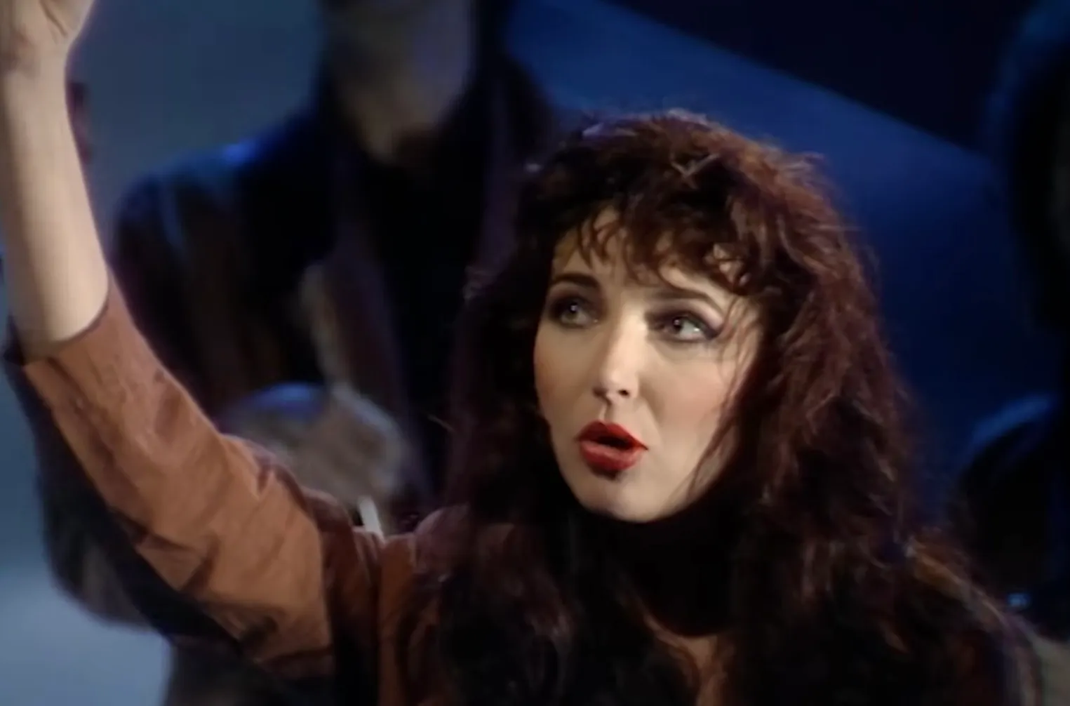 Female 80s celeb known for theatrical performances and hit Running Up That Hill in a dramatic pose with curly hair and red lipstick.
