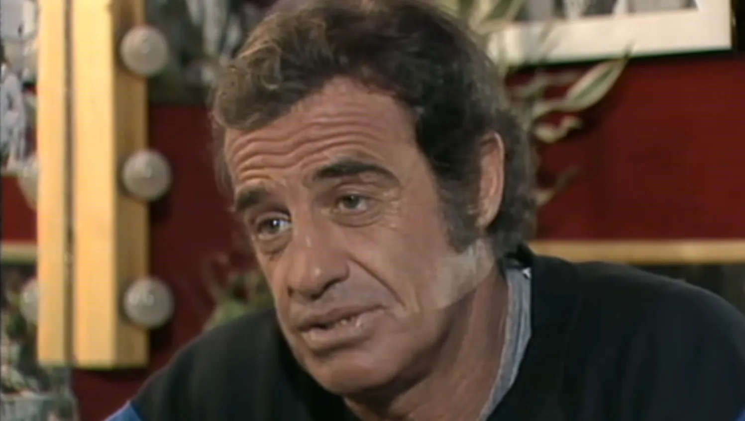 French actor from 80s cinema known for daring stunts and charisma in a close-up interview setting indoors.