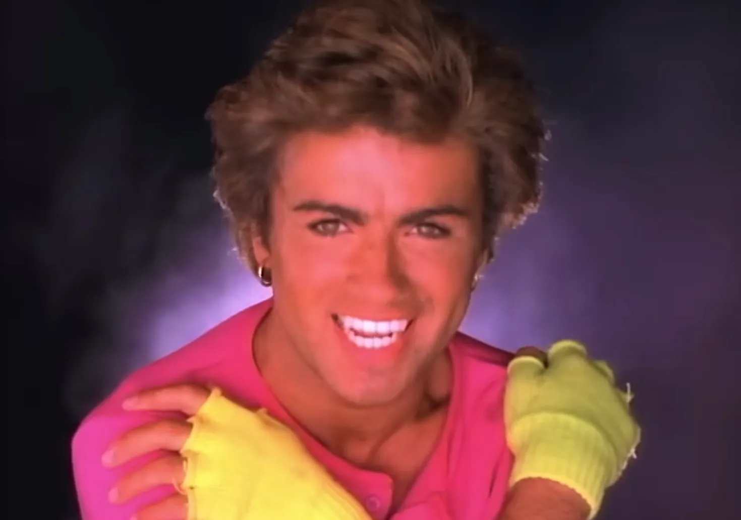 British singer from the 80s wearing bright yellow gloves and a pink top, smiling against a dark smoky background.