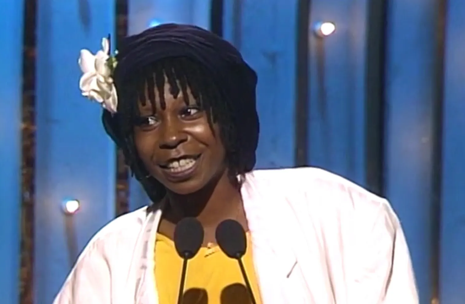 80s celeb at microphone smiling, wearing a white jacket, yellow shirt, black hat with a white flower on stage.