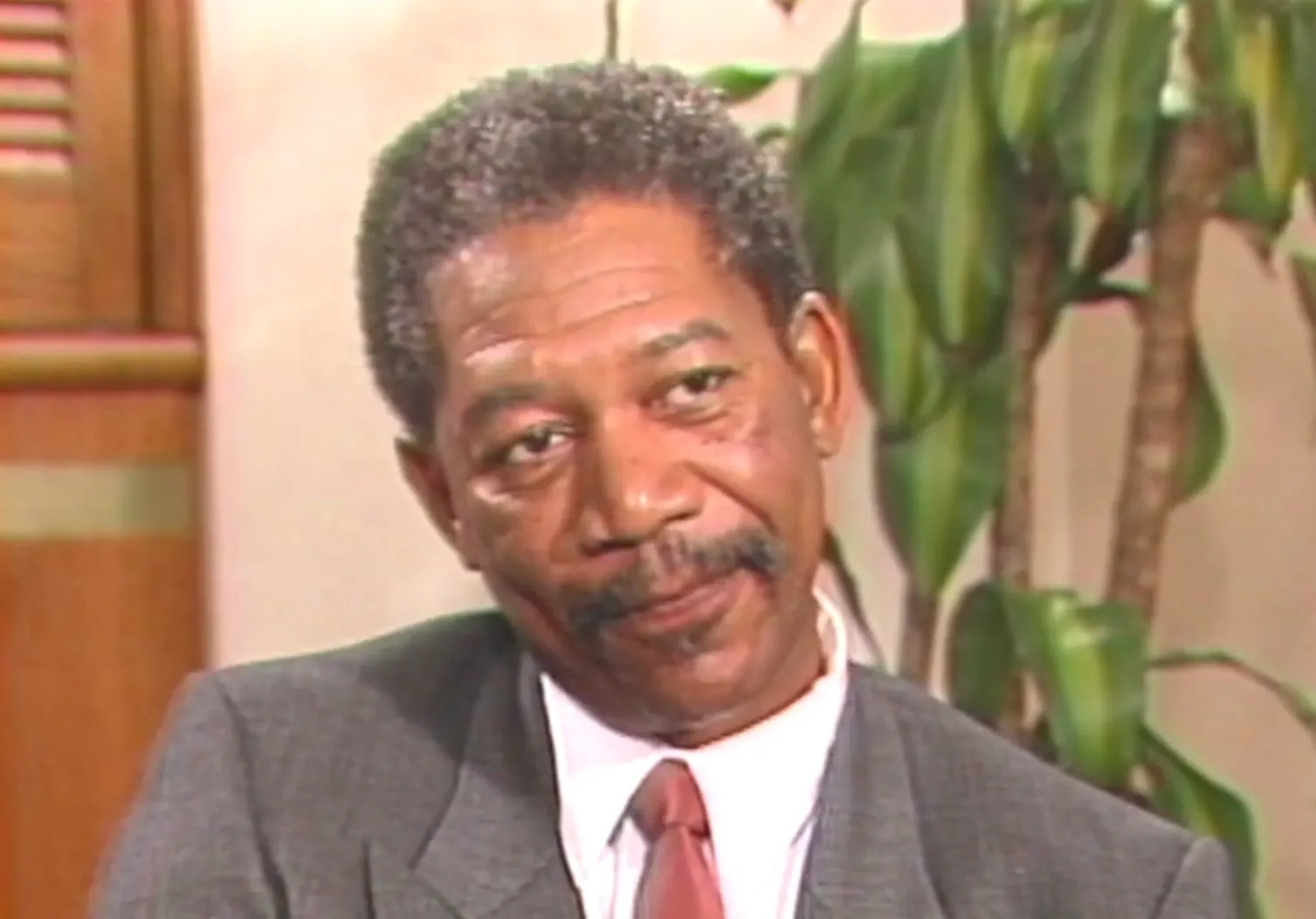 Actor from the 80s in a gray suit and pink tie, sitting indoors with plants and wooden details in the background.