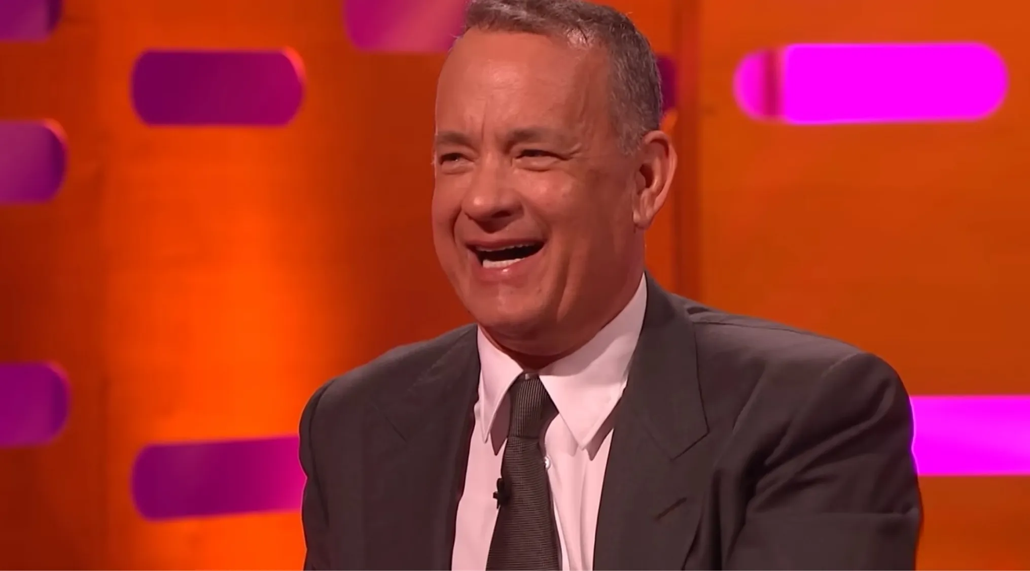 Tom Hanks smiling in a suit and tie during a colorful talk show set guessing movie side characters.