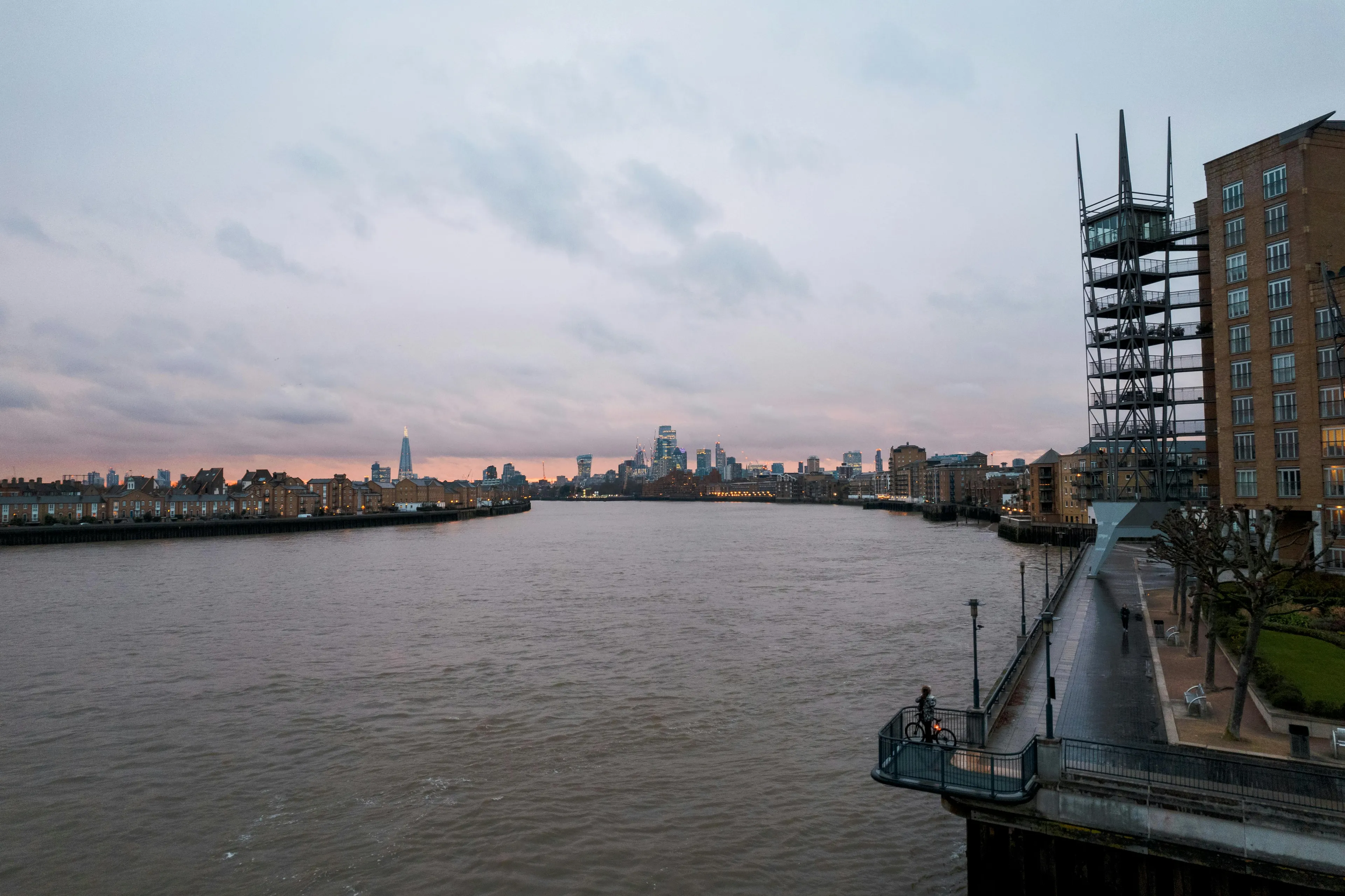 View of the River Thames flowing through the city of London with buildings and a cloudy sky at dusk.
