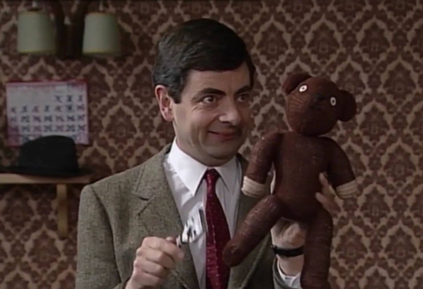 Mr. Bean smiling in a patterned room holding his brown teddy bear, a key character in 80s and 90s TV knowledge.