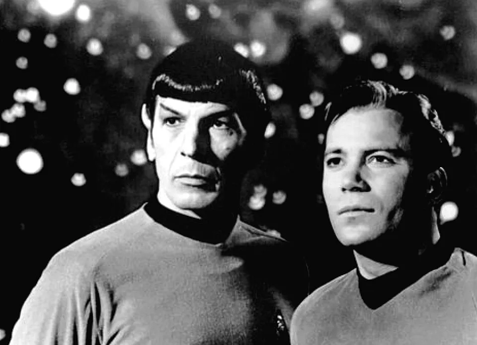 Two male actors from a long-running Star Trek series, wearing classic uniforms, posed in a black and white scene.