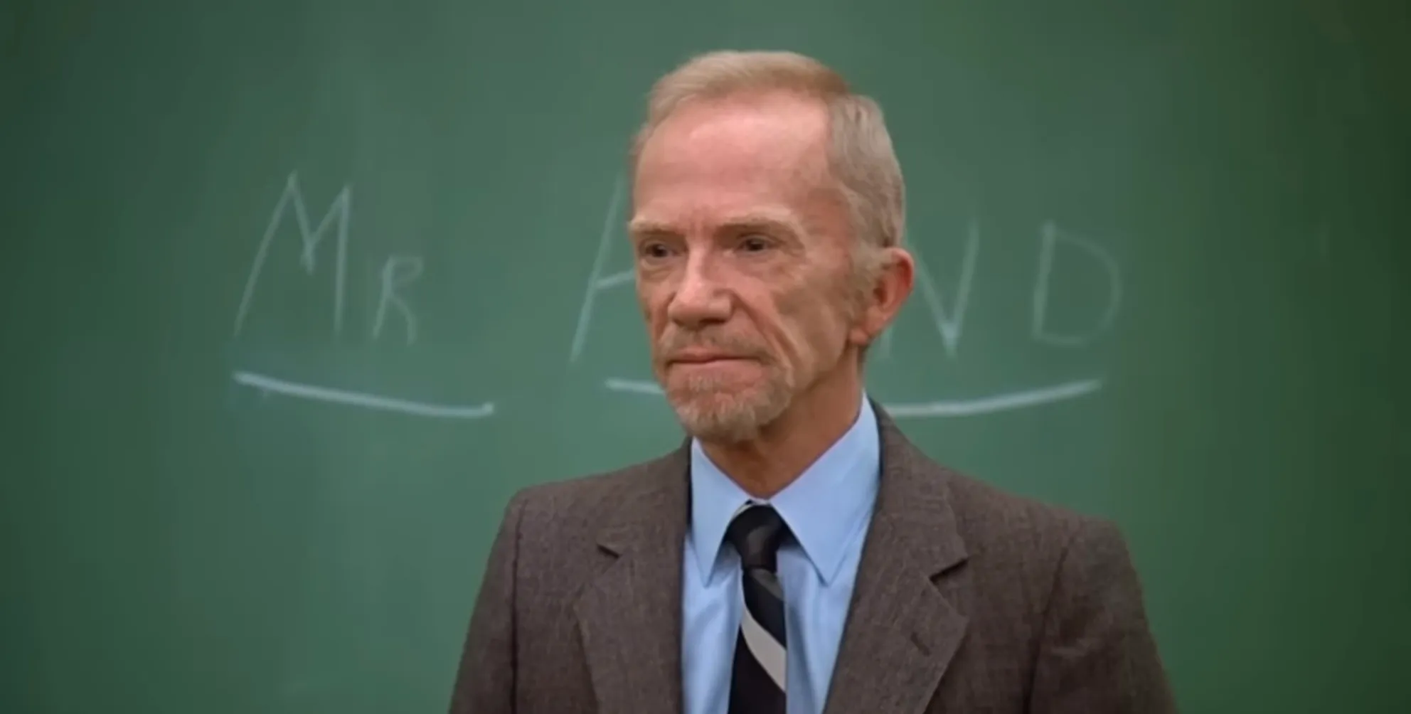 Mr. Hand standing in front of a chalkboard in a brown suit and tie, the principal in Saved by the Bell series.