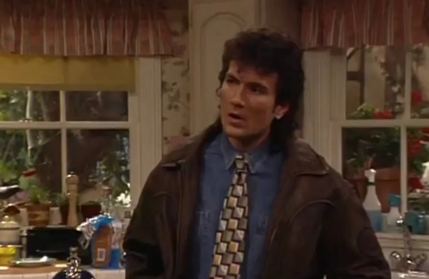 Actor portraying the principal in Saved by the Bell wearing a jacket and patterned tie in a kitchen setting.