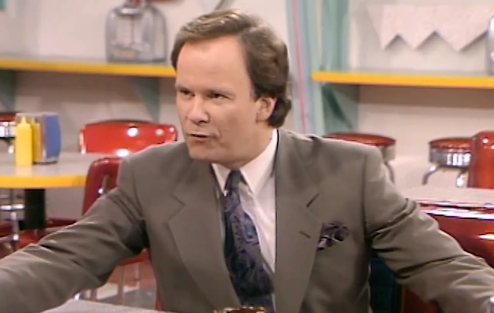Man portraying Mr. Belding, the principal in Saved by the Bell, wearing a suit and sitting in a diner with red chairs.