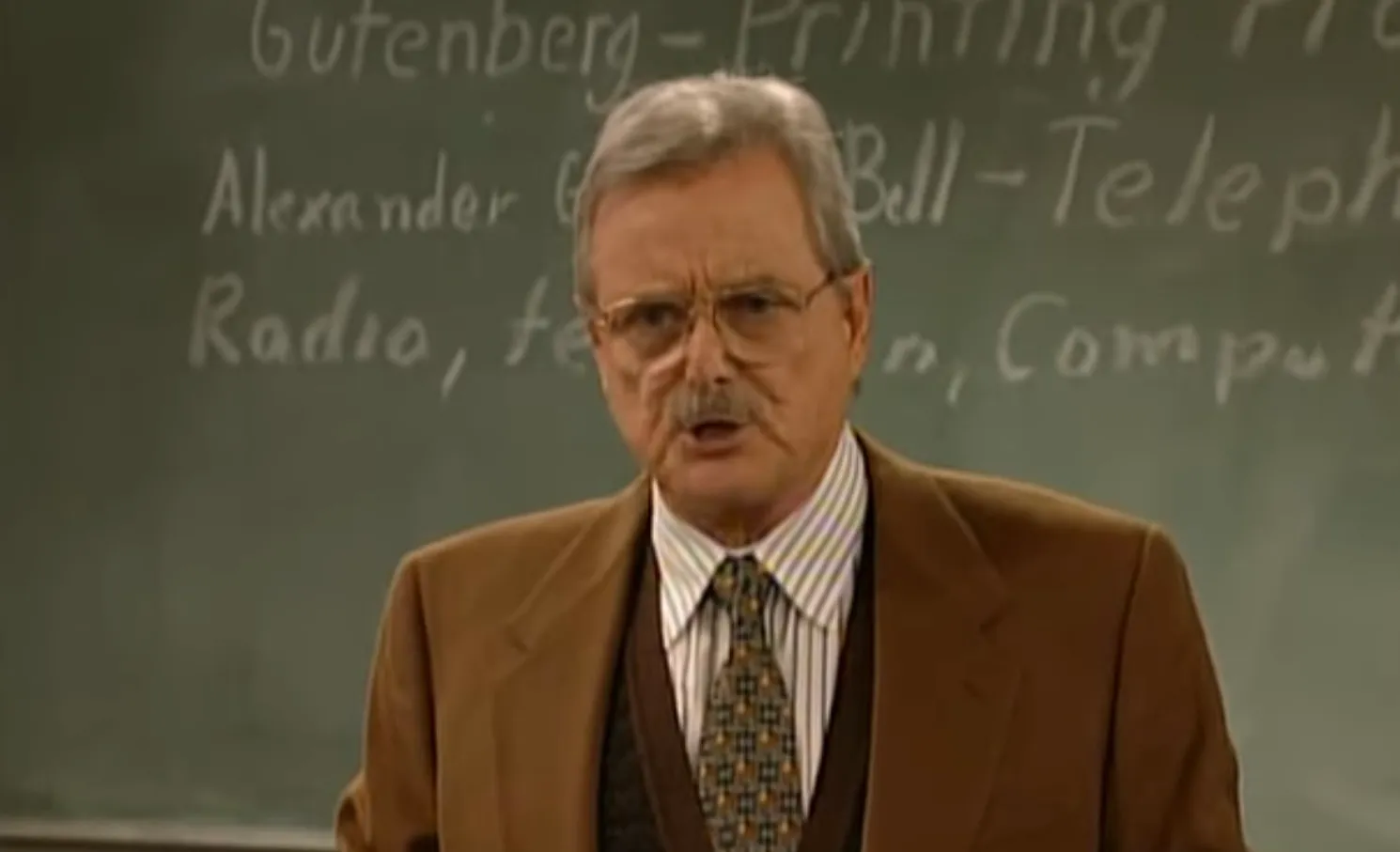 Older man as principal in Saved by the Bell wearing glasses brown suit and standing in front of a classroom chalkboard.