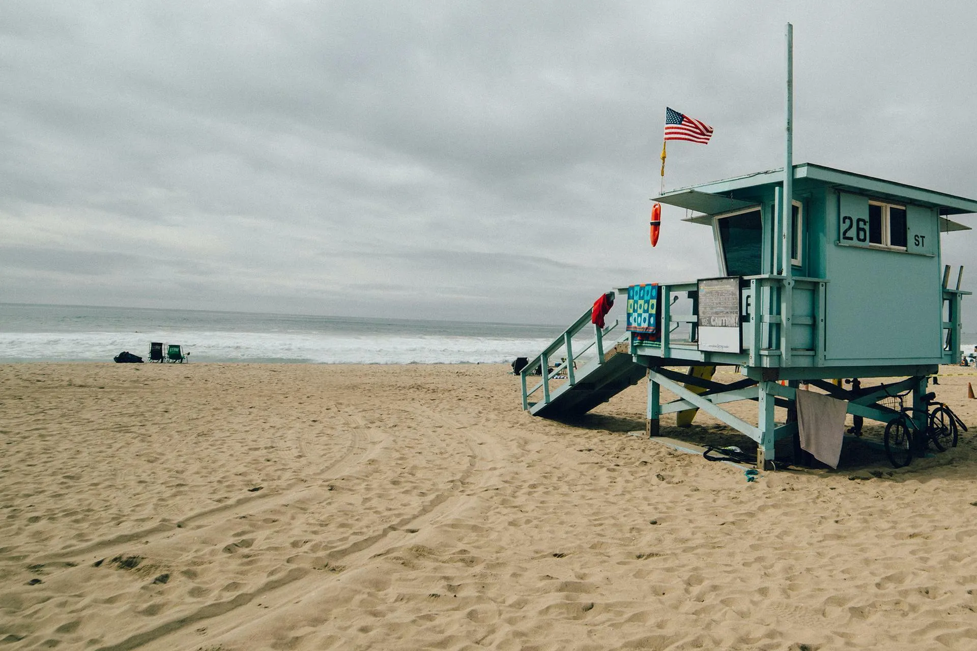 Lifeguard tower on a California beach with an American flag and cloudy sky, representing 80s and 90s TV lifeguards.
