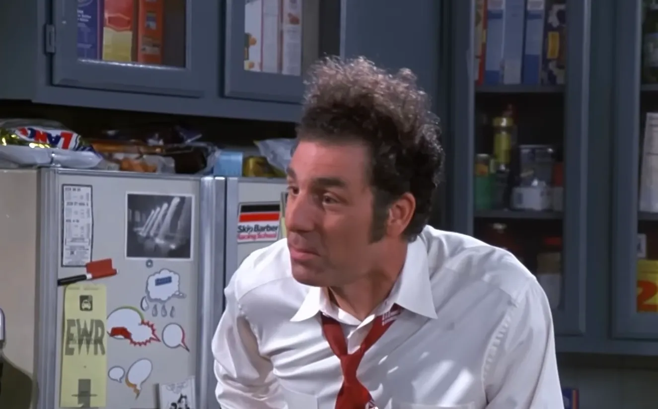 Kramer from Seinfeld in a kitchen scene wearing a white shirt and red tie, showing classic 80s and 90s TV knowledge.