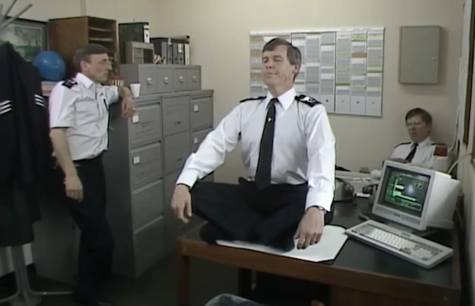 Three officers in uniforms at Sun Hill police station office, one sitting cross-legged on a desk near a vintage computer.