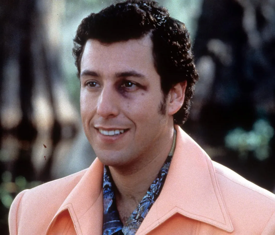 Adam Sandler smiling with a black eye, wearing a peach jacket and patterned shirt in a film scene.