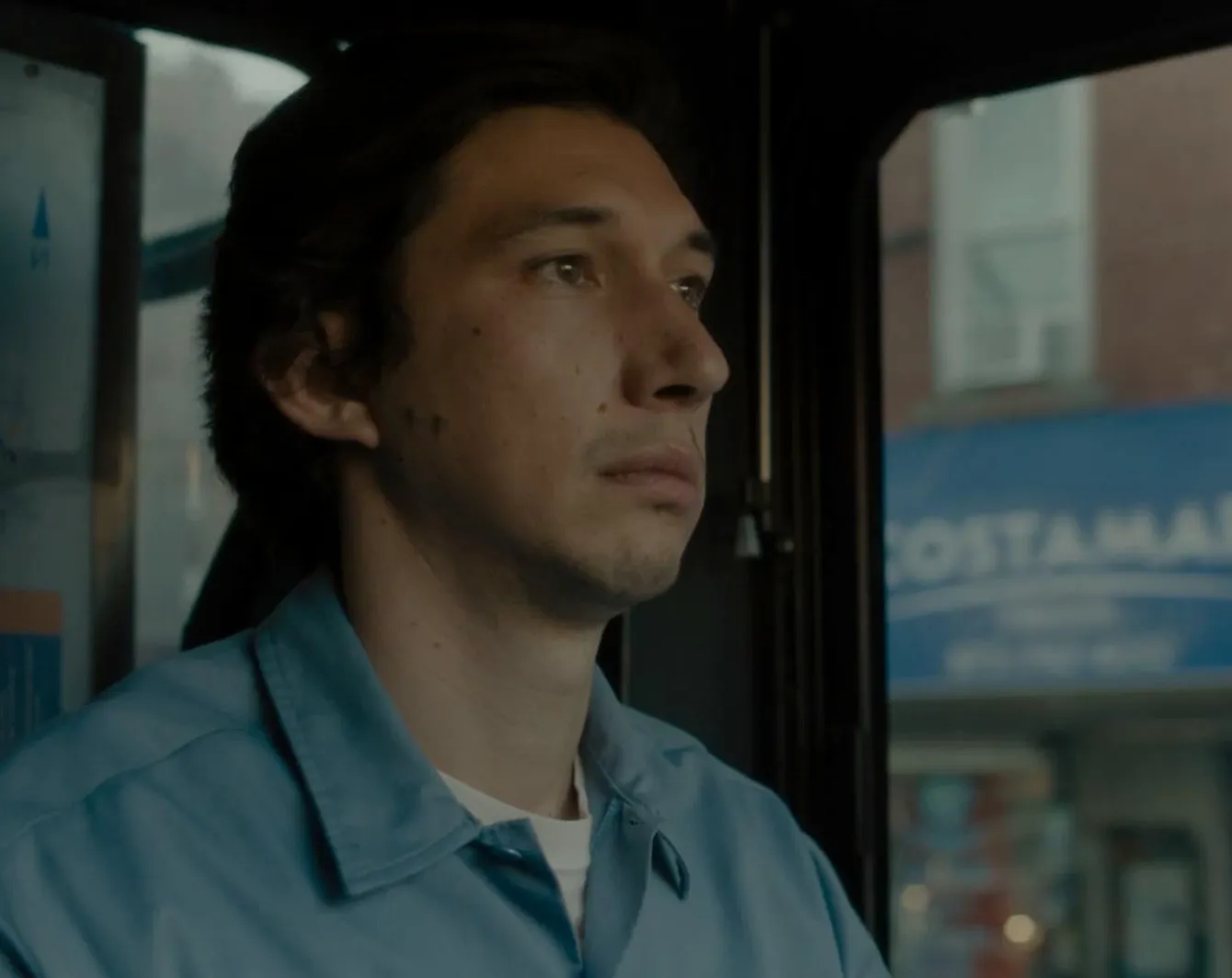 Adam Driver in a scene from Scarface 1983, wearing a blue shirt, looking thoughtfully out of a window.