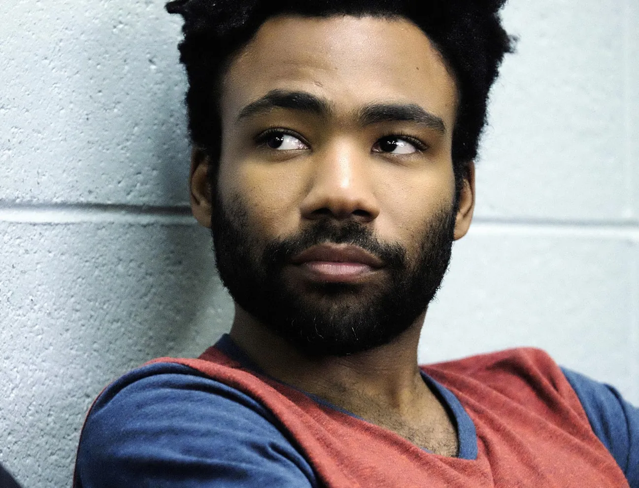 Donald Glover looking thoughtful against a textured wall, wearing a red and blue shirt, actor and musician portrait.
