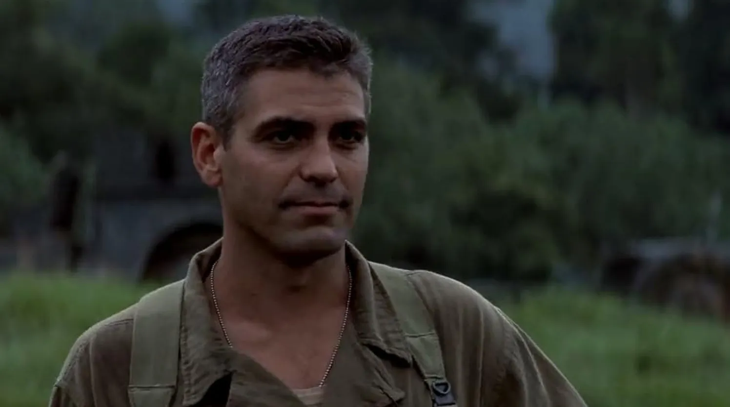 George Clooney in a film scene wearing military attire outdoors with greenery and a blurred background.