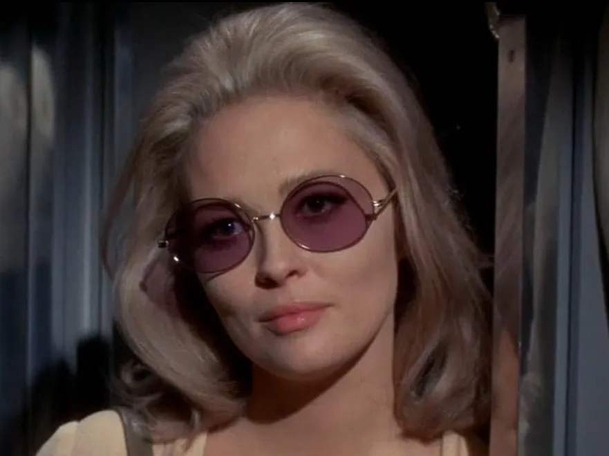 Faye Dunaway wearing round purple sunglasses with light hair, posing indoors in a casual vintage style.