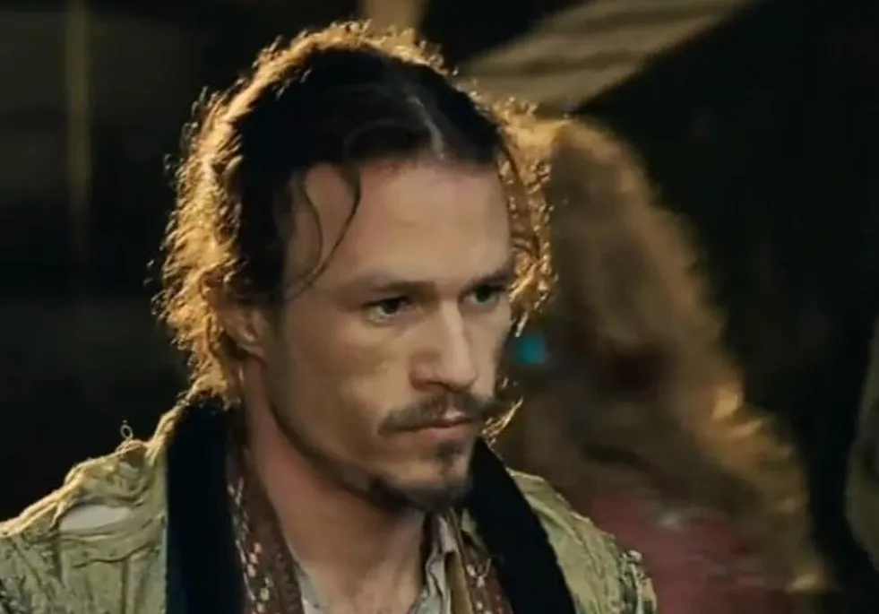 Heath Ledger with curly hair and a mustache in a dimly lit scene, portraying a character from The Dark Knight.