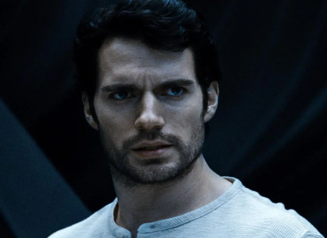 Close-up of Henry Cavill with dark hair and a beard, wearing a light shirt in a dimly lit setting.