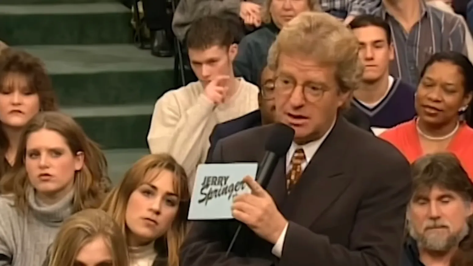 Jerry Springer hosting with microphone and audience behind, illustrating 90s TV knowledge and show format.