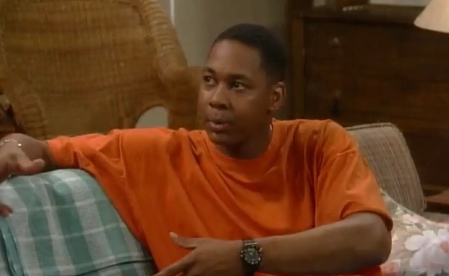 Man in an orange shirt sitting on a couch in a living room, representing 80s and 90s TV knowledge Hangin with Mr Cooper focus.