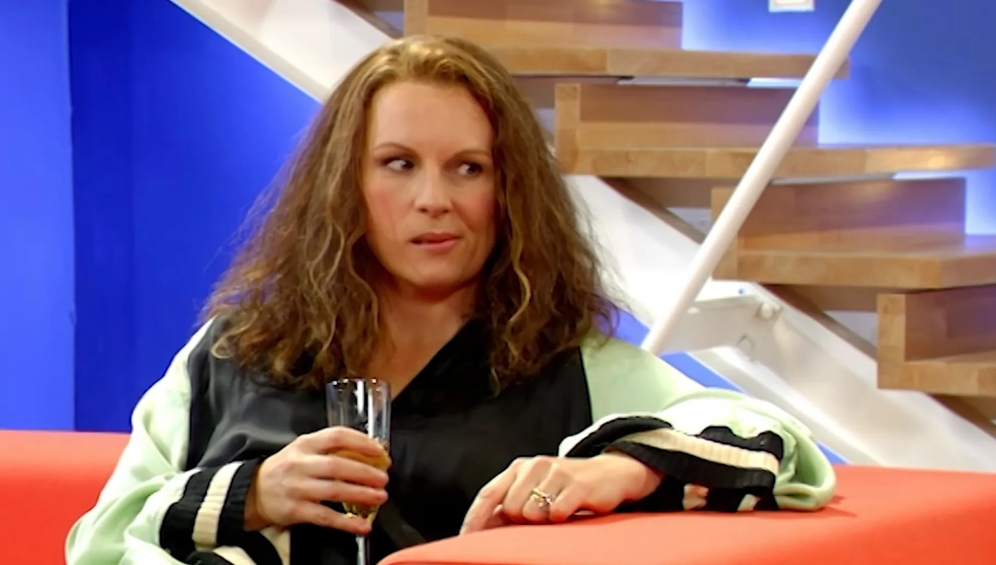 Woman with curly hair holding a glass sitting on an orange couch, related to Edina Monsoon’s profession in 80s and 90s TV.