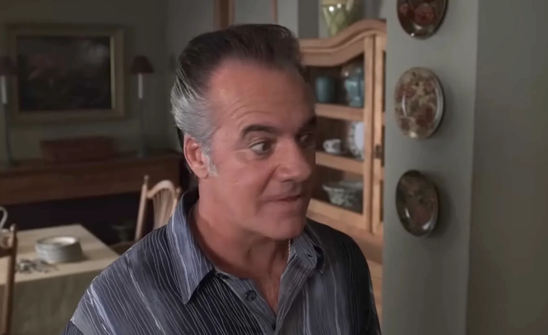 Paulie Walnuts from The Sopranos in a striped shirt, standing indoors with plates on the wall behind him.