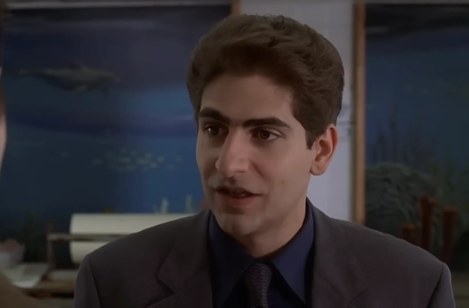 Christopher Moltisanti from The Sopranos wearing a dark suit and tie, engaged in conversation indoors with ocean-themed decor.