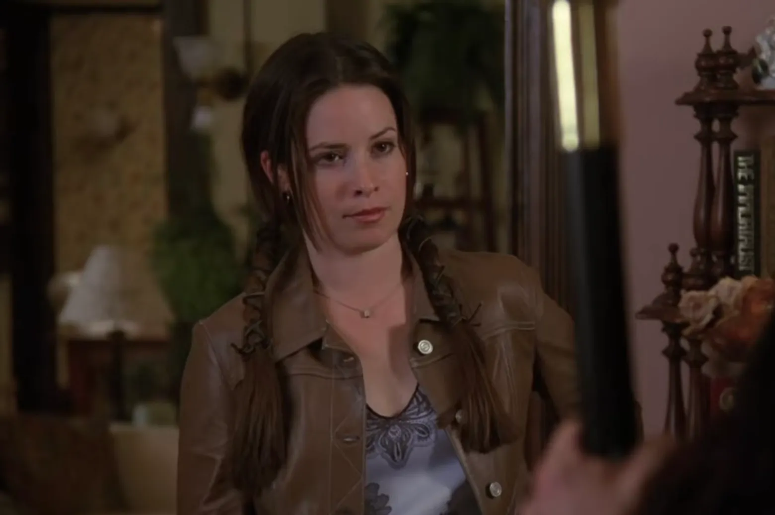 Piper Halliwell from Charmed wearing a brown jacket and standing indoors with braided hair and a serious expression.
