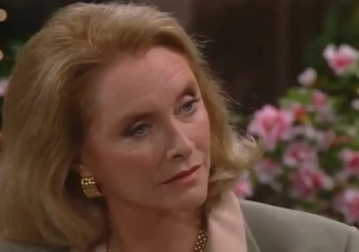 Stephanie Forrester from The Bold and the Beautiful with blonde hair, wearing gold earrings, in an indoor setting with flowers.