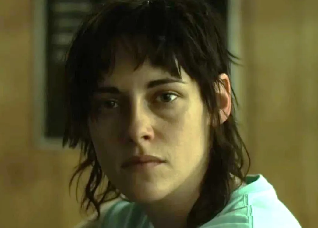 Kristen Stewart with short dark hair, wearing a light-colored shirt in a dimly lit indoor setting.