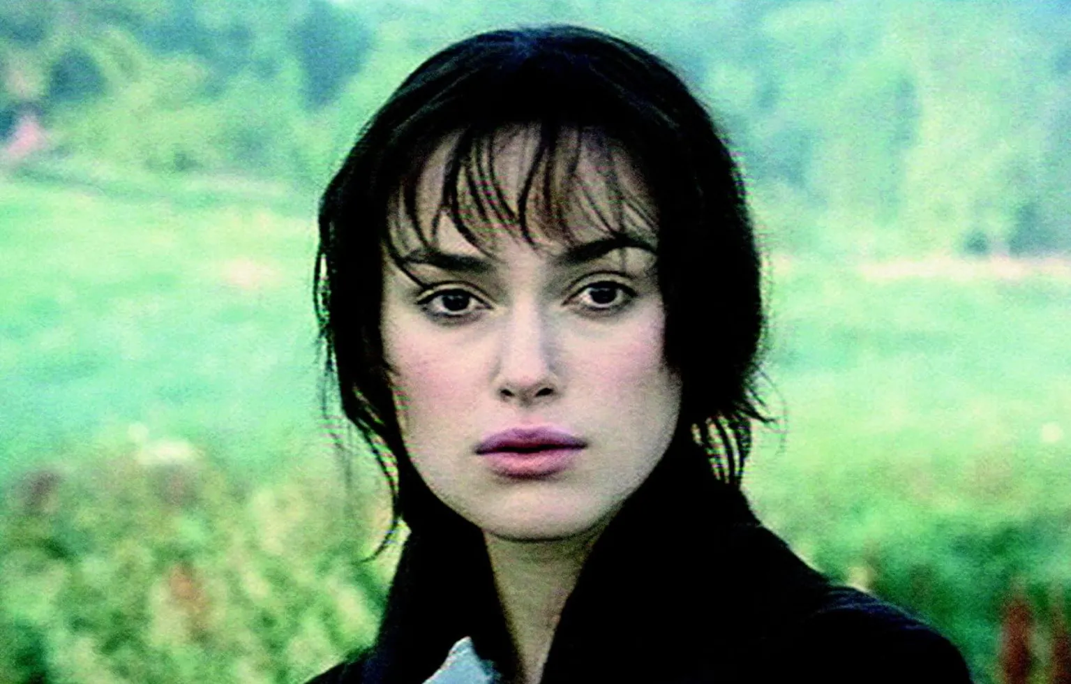 Close-up of actress Keira Knightley with dark hair and bangs, outdoors with a green blurred background.