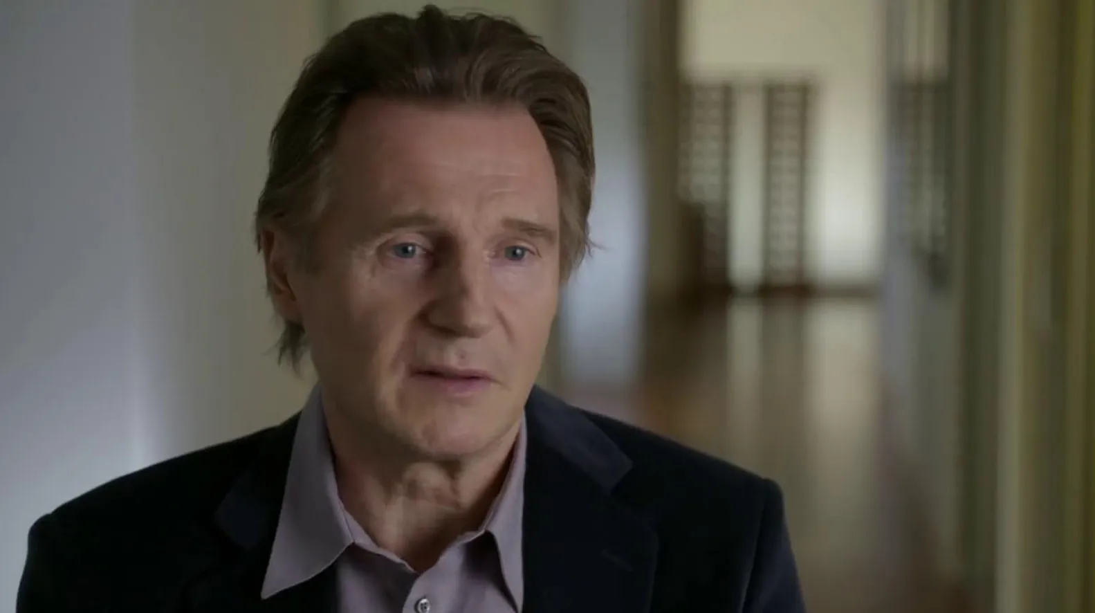 Liam Neeson in a close-up interview shot, wearing a dark jacket and light shirt, with a blurred hallway background.