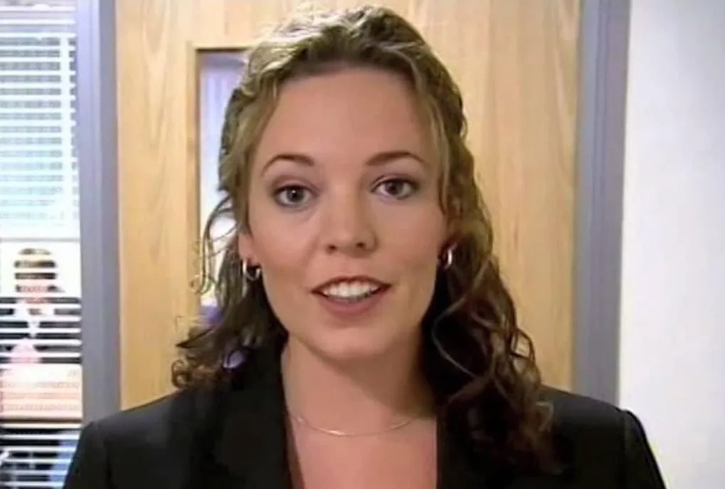 Actress Olivia Colman smiling indoors, with curly hair, wearing a black blazer in an office setting.