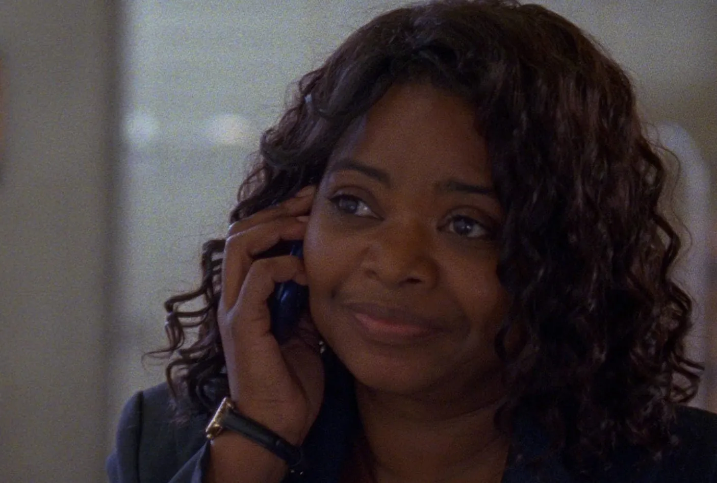 Octavia Spencer talking on phone, smiling gently, with curly hair and wearing a dark jacket in an indoor setting.