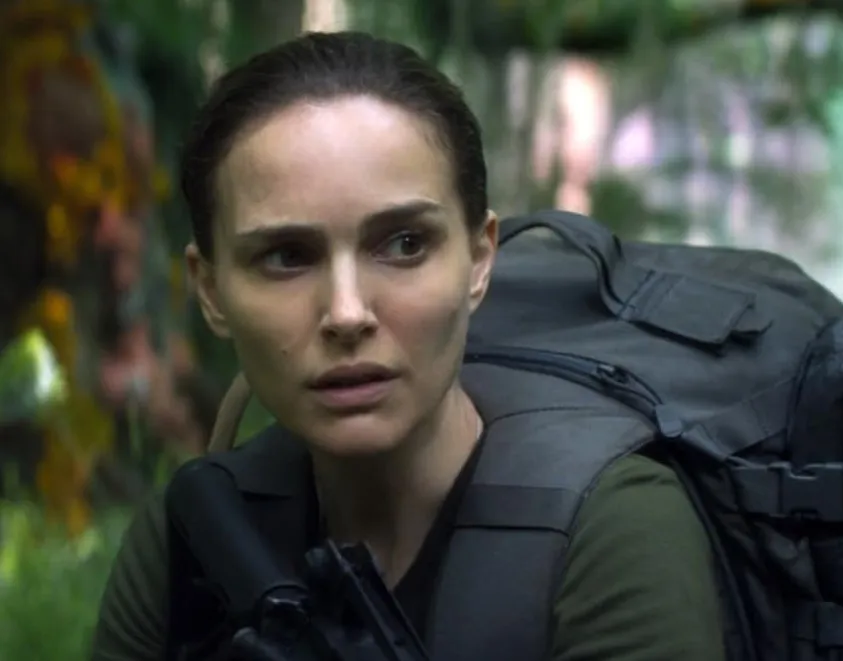Natalie Portman in a tense scene wearing tactical gear and carrying a backpack in a forested outdoor setting.