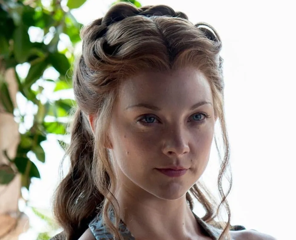 Close-up of actress Natalie Dormer with braided hair and a calm expression, surrounded by natural light and greenery.