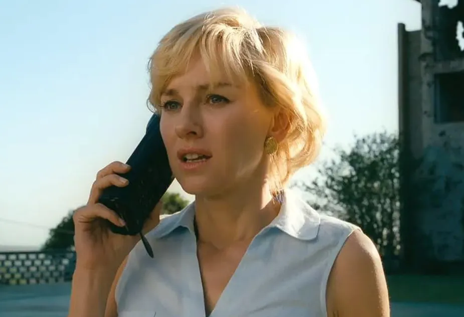 Naomi Watts in a sleeveless top speaking on a phone outdoors with a blurred background and natural lighting.
