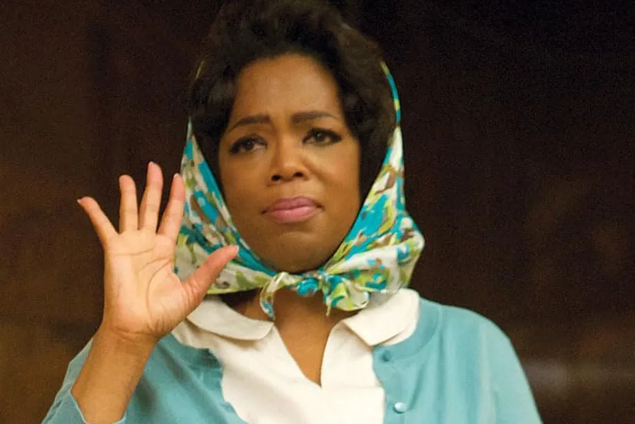 Oprah Winfrey wearing a patterned headscarf and blue cardigan, raising her hand in an emotional scene